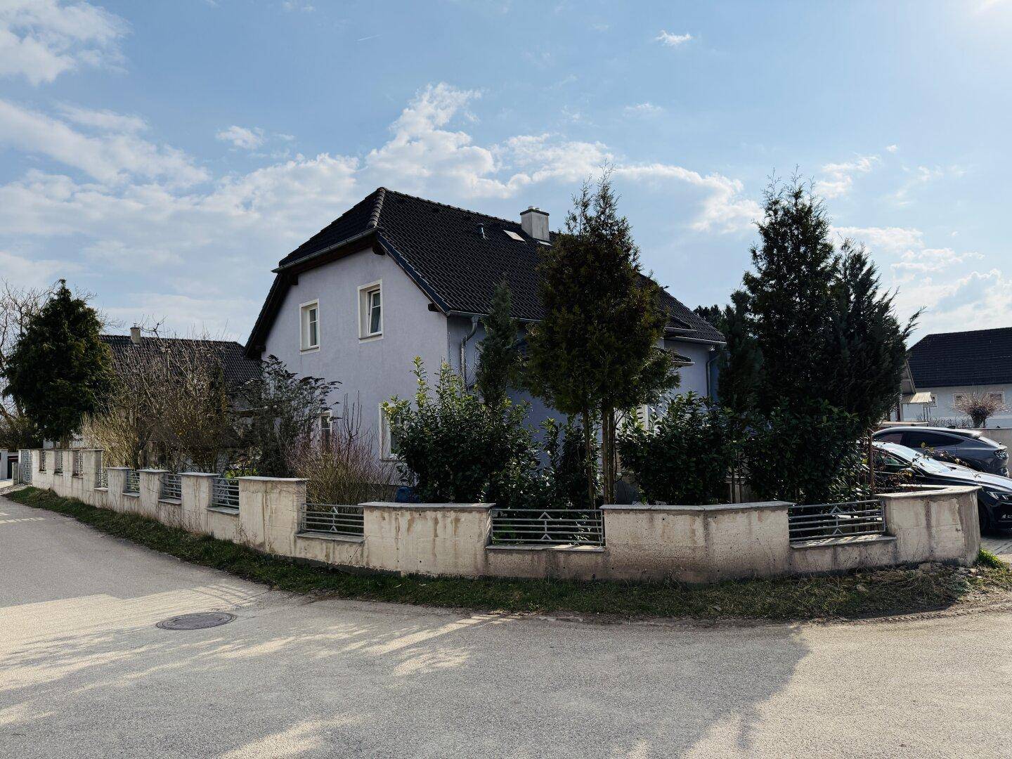 Seitenansicht des Hauses von der Straße aus, umgeben von einer niedrigen Mauer und Bäumen.