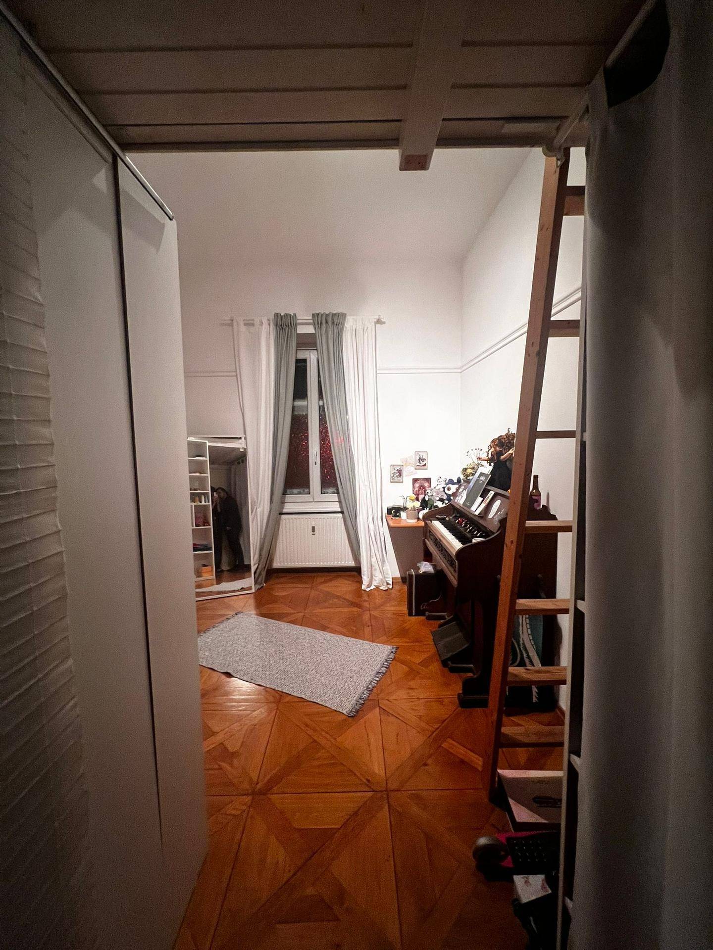 Zimmer mit Holzboden, Fenster und einer Leiter, die zu einem Hochbett oder Stauraum führt.