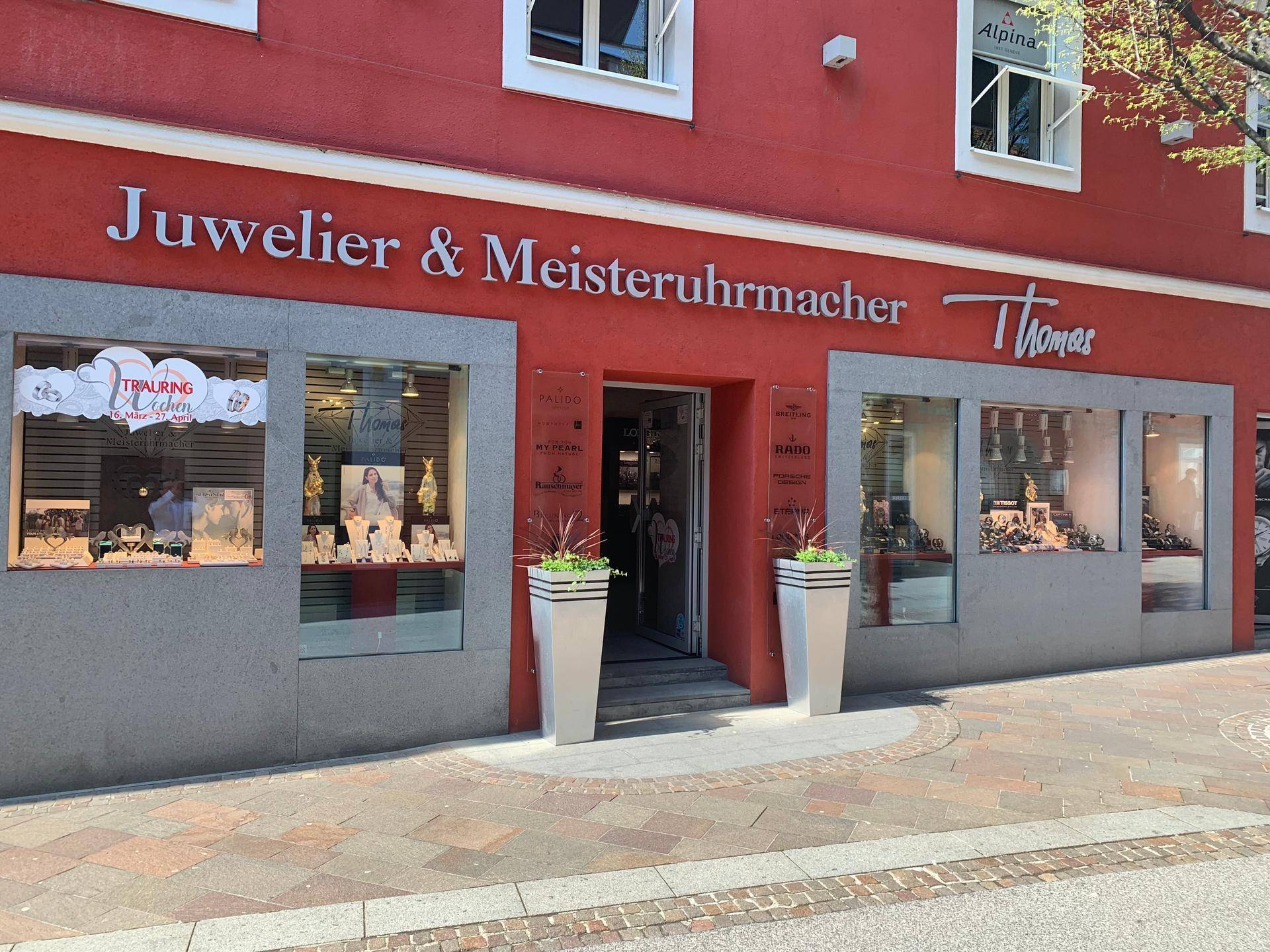 Ansicht der roten Fassade eines Juweliergeschäfts mit hellen Schaufenstern, die Uhren und Schmuck präsentieren.
