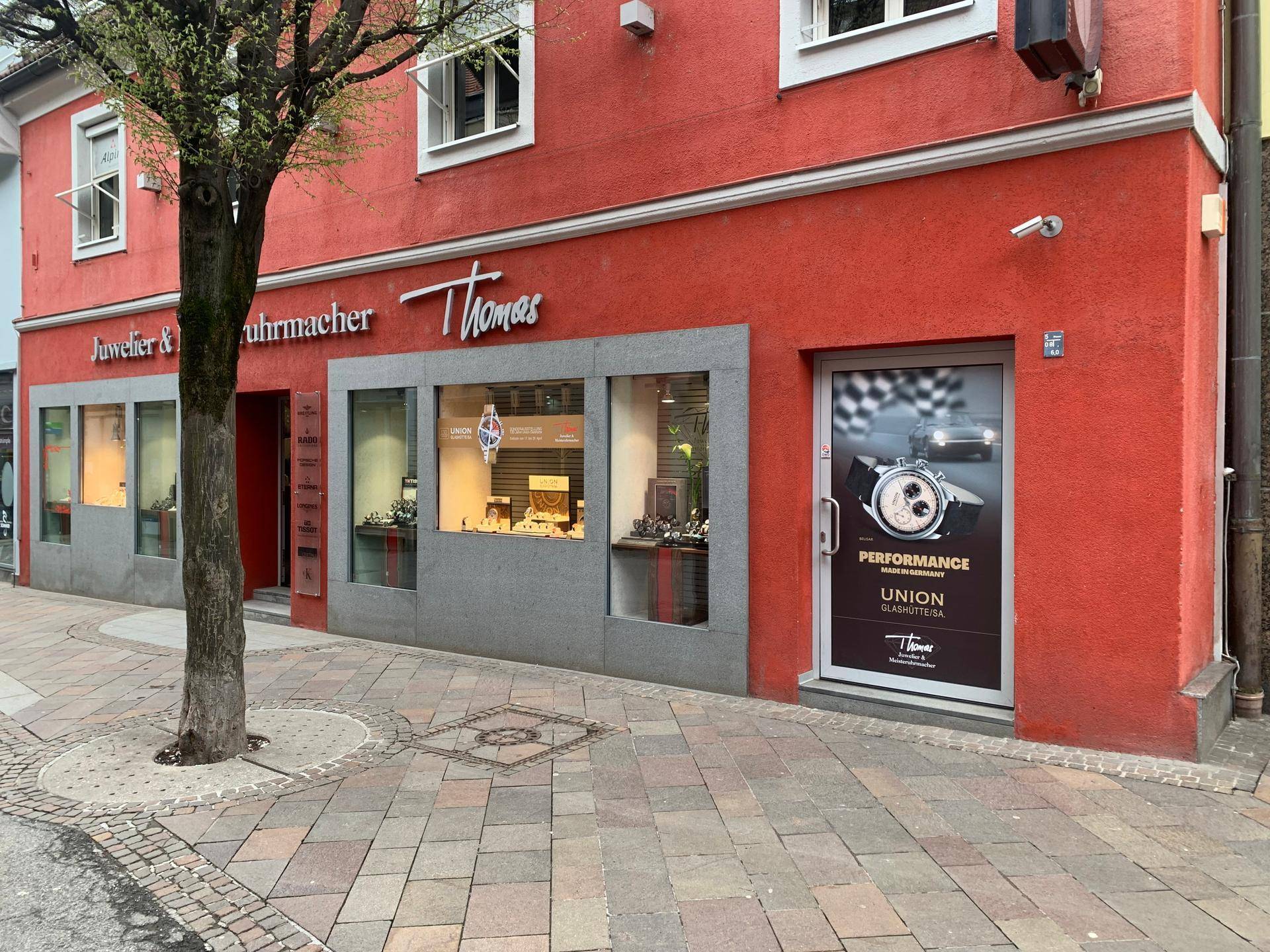 Geschäftsfassade mit roten Wänden, großen Schaufenstern und einem Baum im Vordergrund am Bürgersteig.