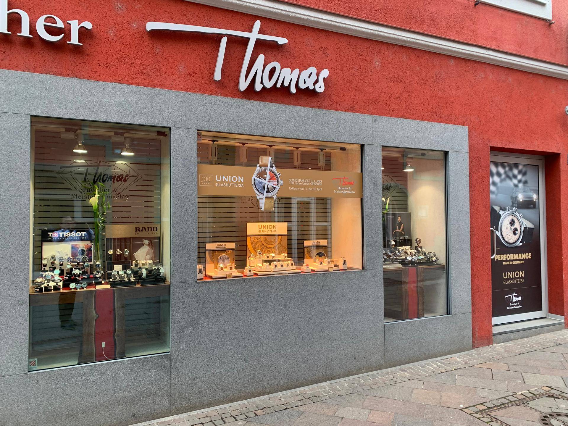 Nahaufnahme der rechten Schaufenster eines Uhrmachergeschäfts mit Uhrenauslagen und einer Werbetür.