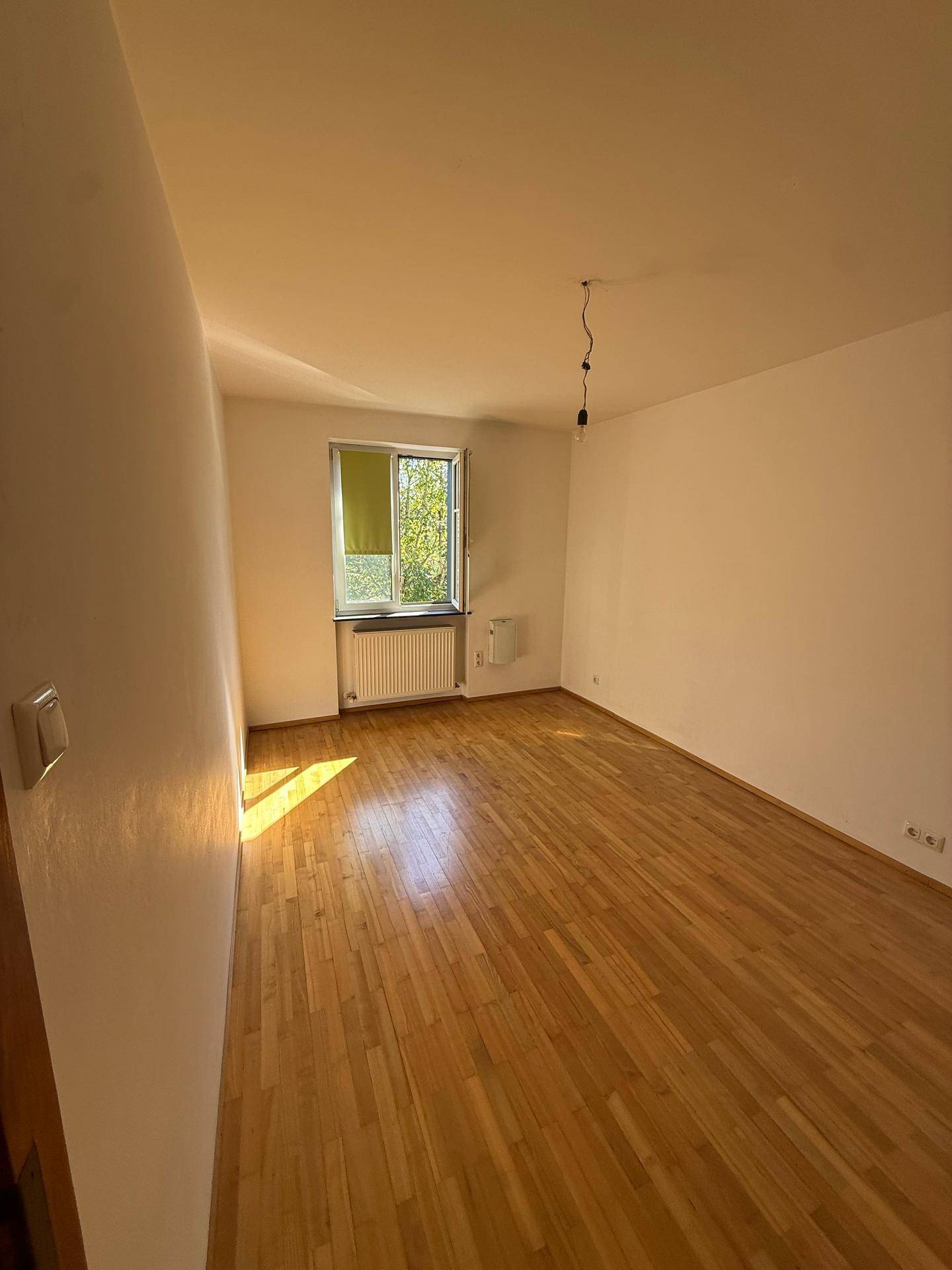 Helles Schlafzimmer mit Holzboden, weißen Wänden und einem Fenster, durch das Sonnenlicht einfällt.