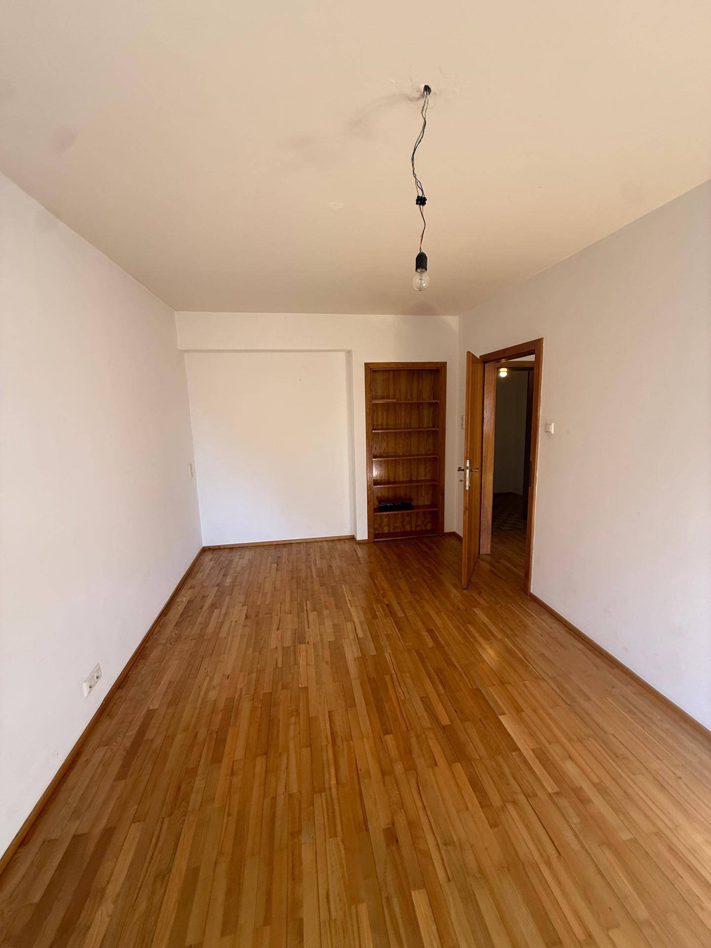 Leeres Zimmer mit hellem Holzboden, weißen Wänden und einem eingebauten Holzregal.