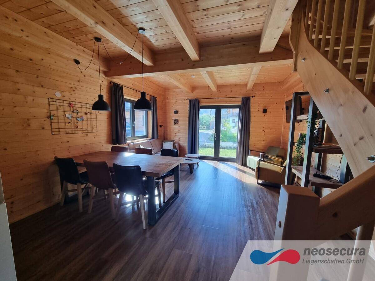 Lichtdurchfluteter Wohn-Essbereich mit viel Holz, großen Fenstern und Blick auf die grüne Umgebung.