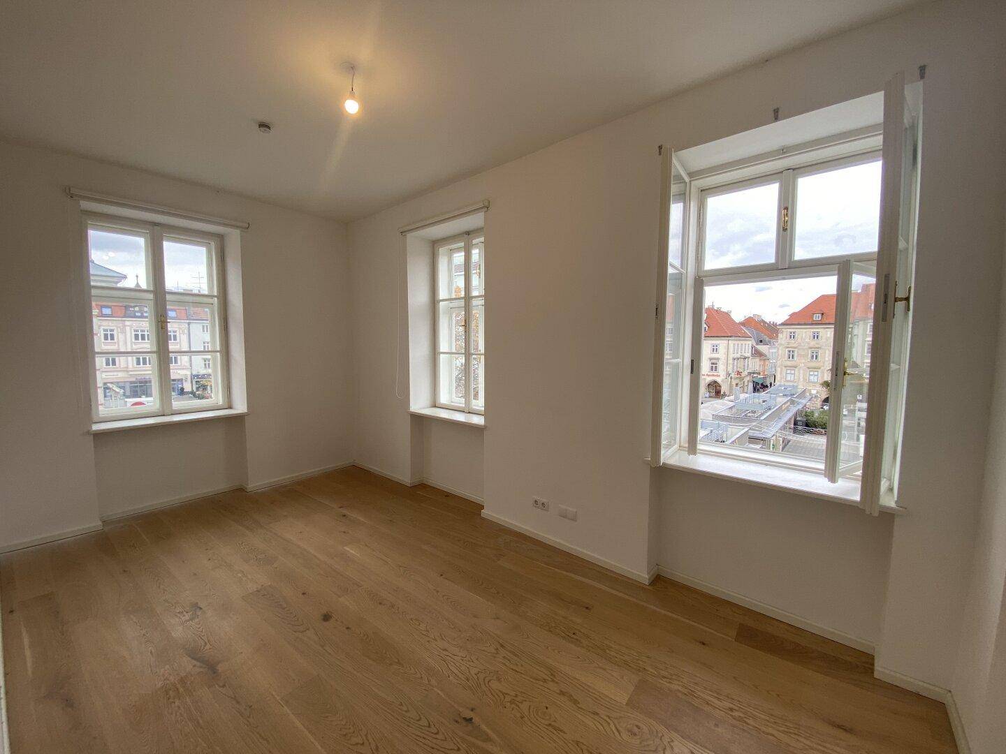 Geräumiges Wohnzimmer mit drei großen Fenstern und hellem Parkettboden, das viel Licht bietet.