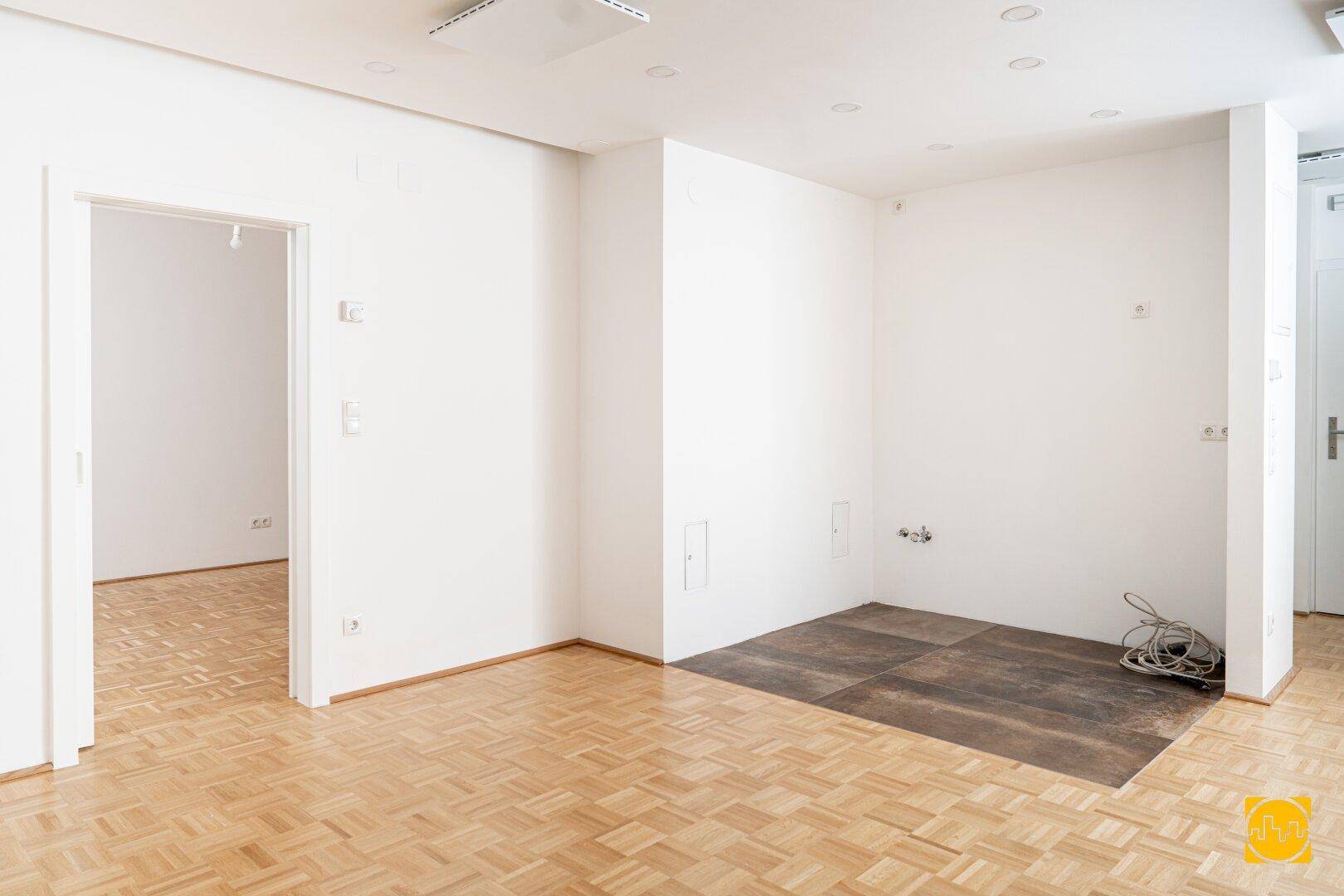 Heller Raum mit Parkettboden und vorbereitetem Bereich für eine moderne Küche.