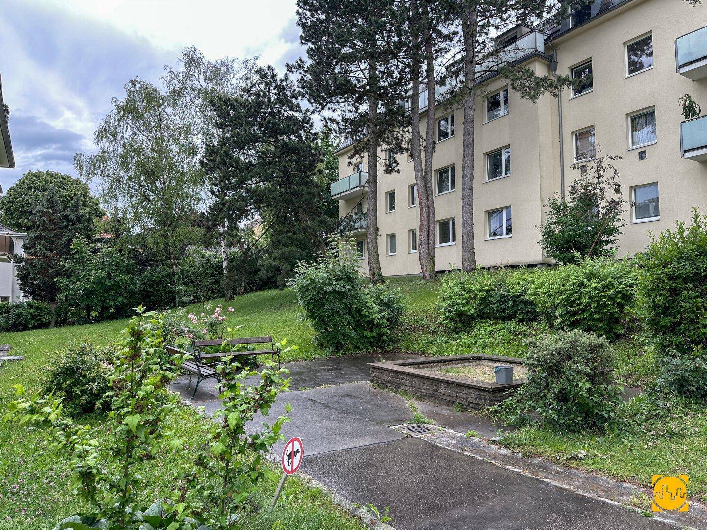Gepflegter Gemeinschaftsgarten mit Sitzbank, Sandkasten und altem Baumbestand vor dem Gebäude.