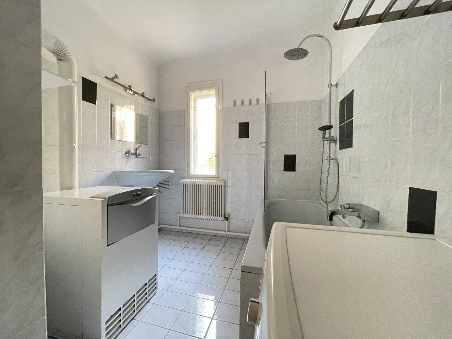 Helles Badezimmer mit Badewanne, Dusche und Fenster, ausgestattet mit weißen Fliesen.