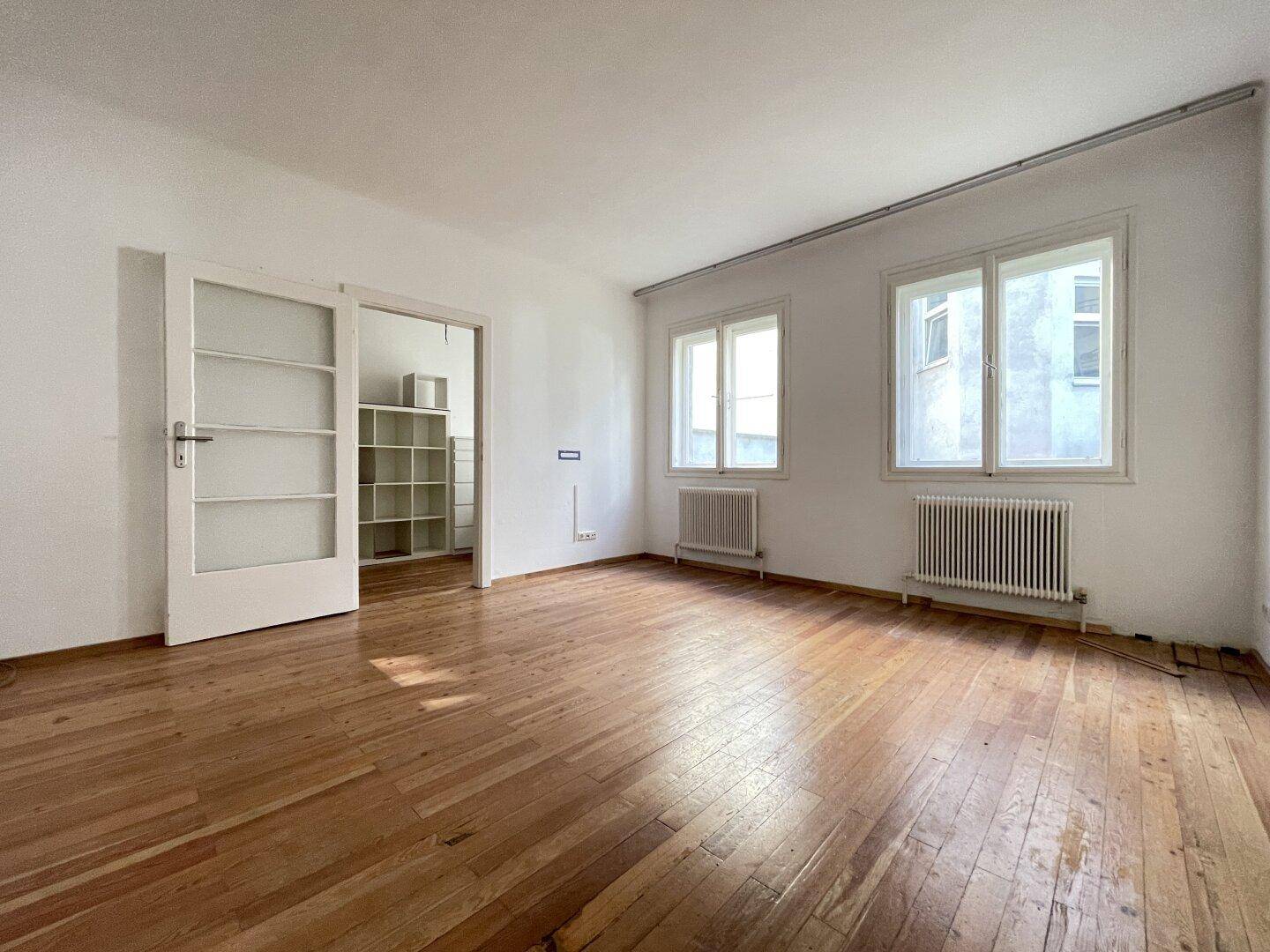 Geräumiges Wohnzimmer mit Holzboden, weißen Wänden und zwei großen Fenstern.