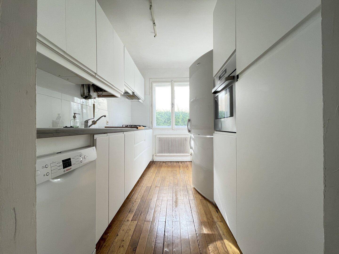 Schmale, moderne Küche mit weißen Schränken, Holzboden und Blick ins Grüne durch das Fenster.