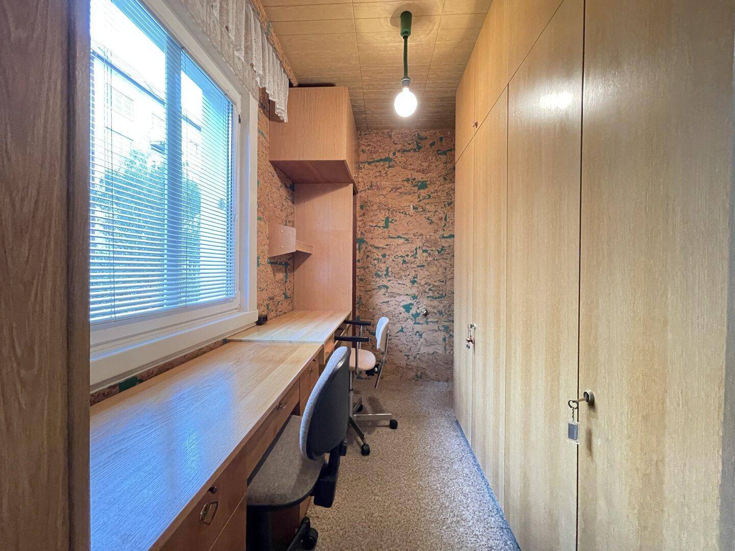 Schmaler Arbeitsraum mit langen Schreibtischen, Einbauschränken und Fenster.