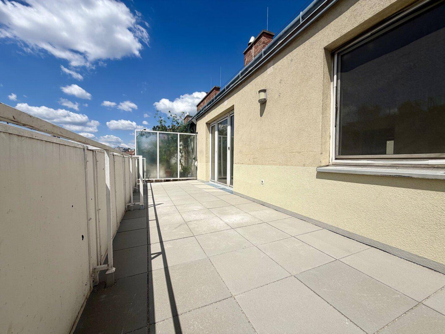 Geräumige Dachterrasse mit Fliesenboden und Glasgeländer unter blauem Himmel.