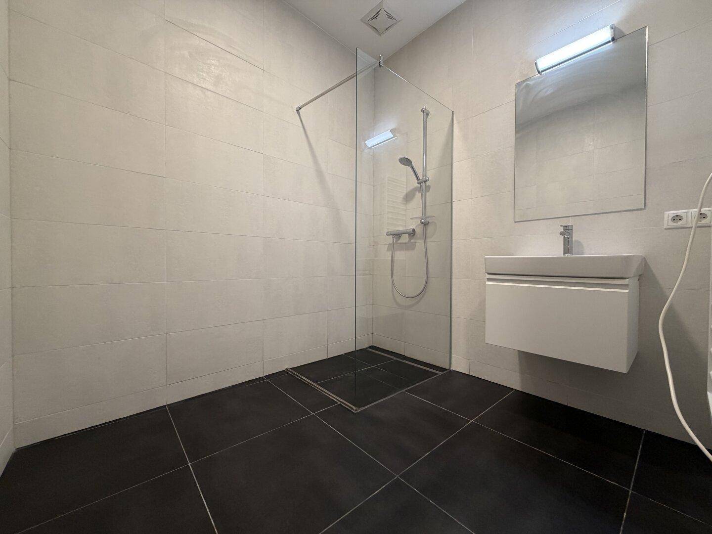 Modernes Badezimmer mit dunklen Bodenfliesen, begehbarer Dusche und wandmontiertem Waschbecken mit Spiegel.