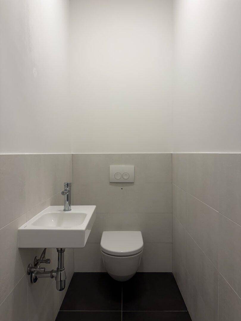 Modernes Gäste-WC mit wandhängender Toilette, kleinem Waschbecken und dunklen Bodenfliesen.
