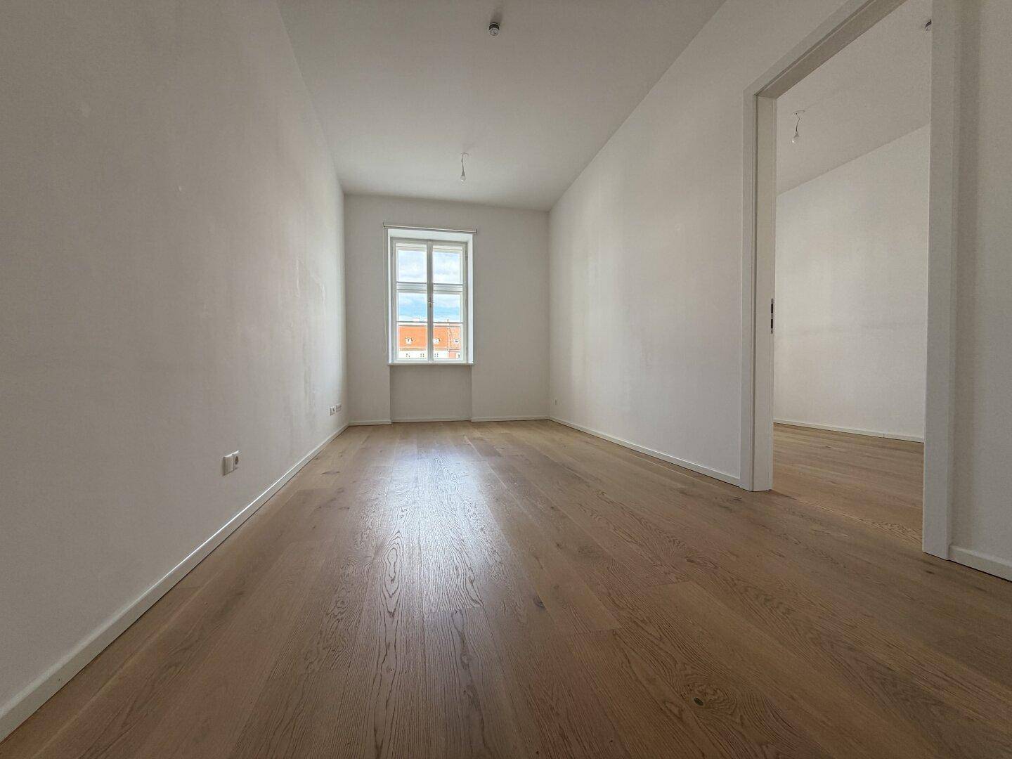Heller, leerer Raum mit neuem Holzparkettboden und einem Fenster, das viel Tageslicht hereinlässt.