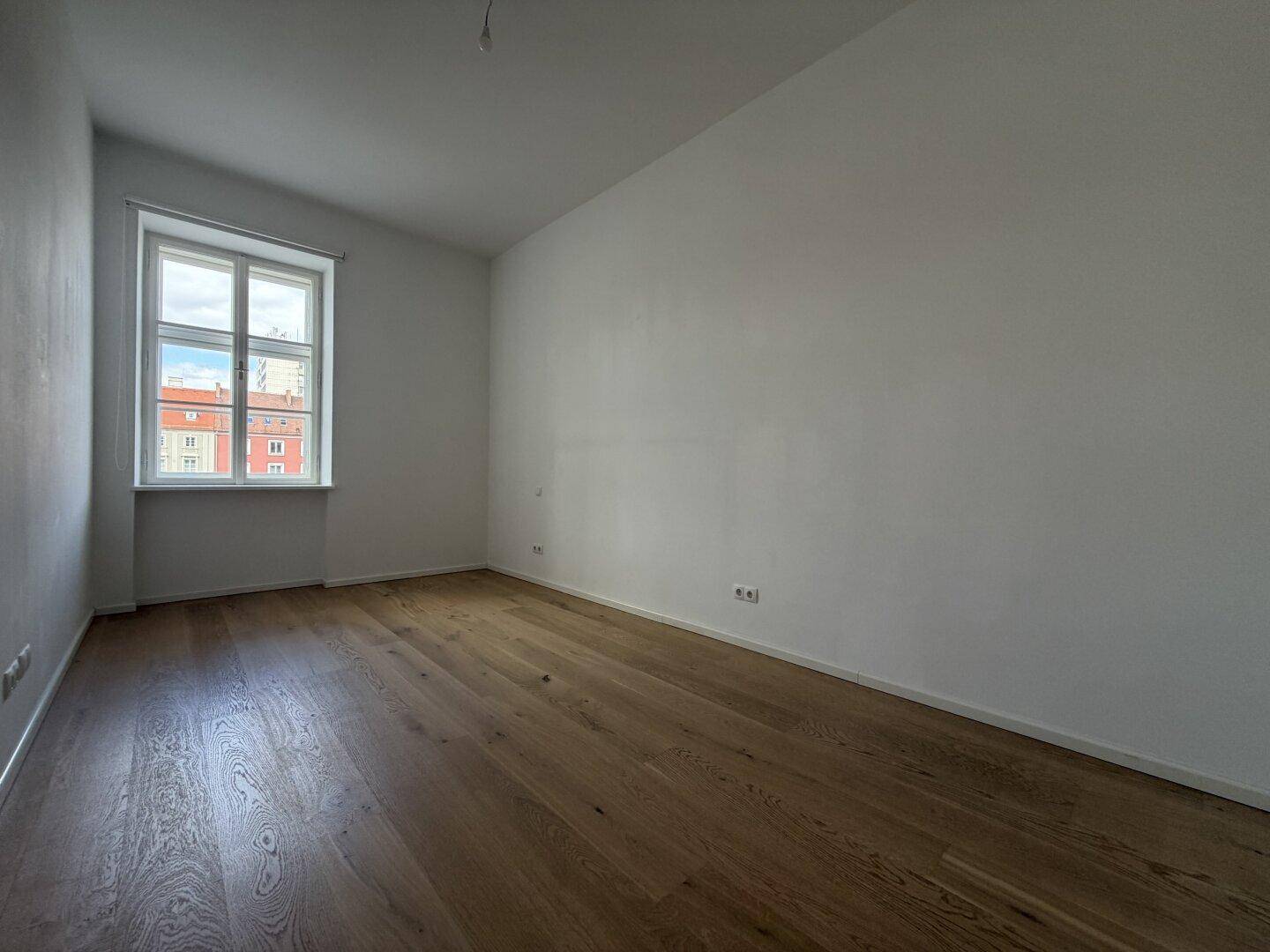 Geräumiges, helles Zimmer mit neuem Holzparkettboden und einem Fenster, ideal als Schlafzimmer nutzbar.