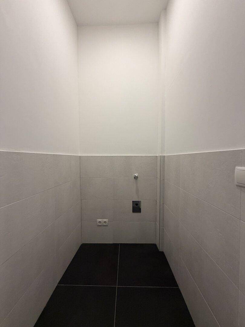Schmaler Raum mit weißen Wänden und grauen Fliesen, vorbereitet für eine Toilette oder Waschküche.