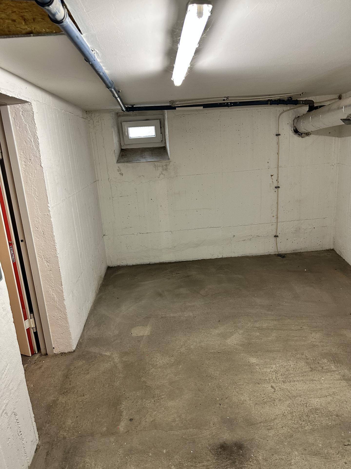 Leerer Kellerraum mit Betonboden, weiß gestrichenen Wänden und einem kleinen Fenster.