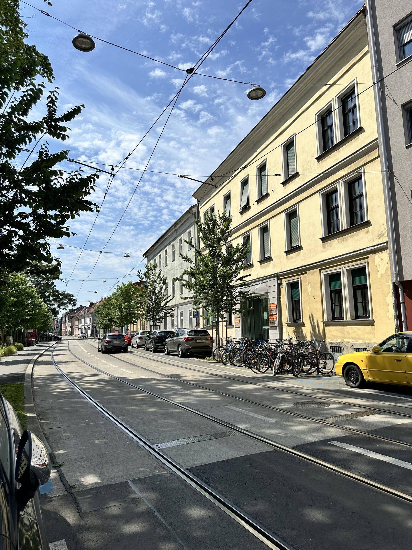 Eine belebte Straße mit Straßenbahnschienen, parkenden Autos und Bäumen unter einem sonnigen Himmel.