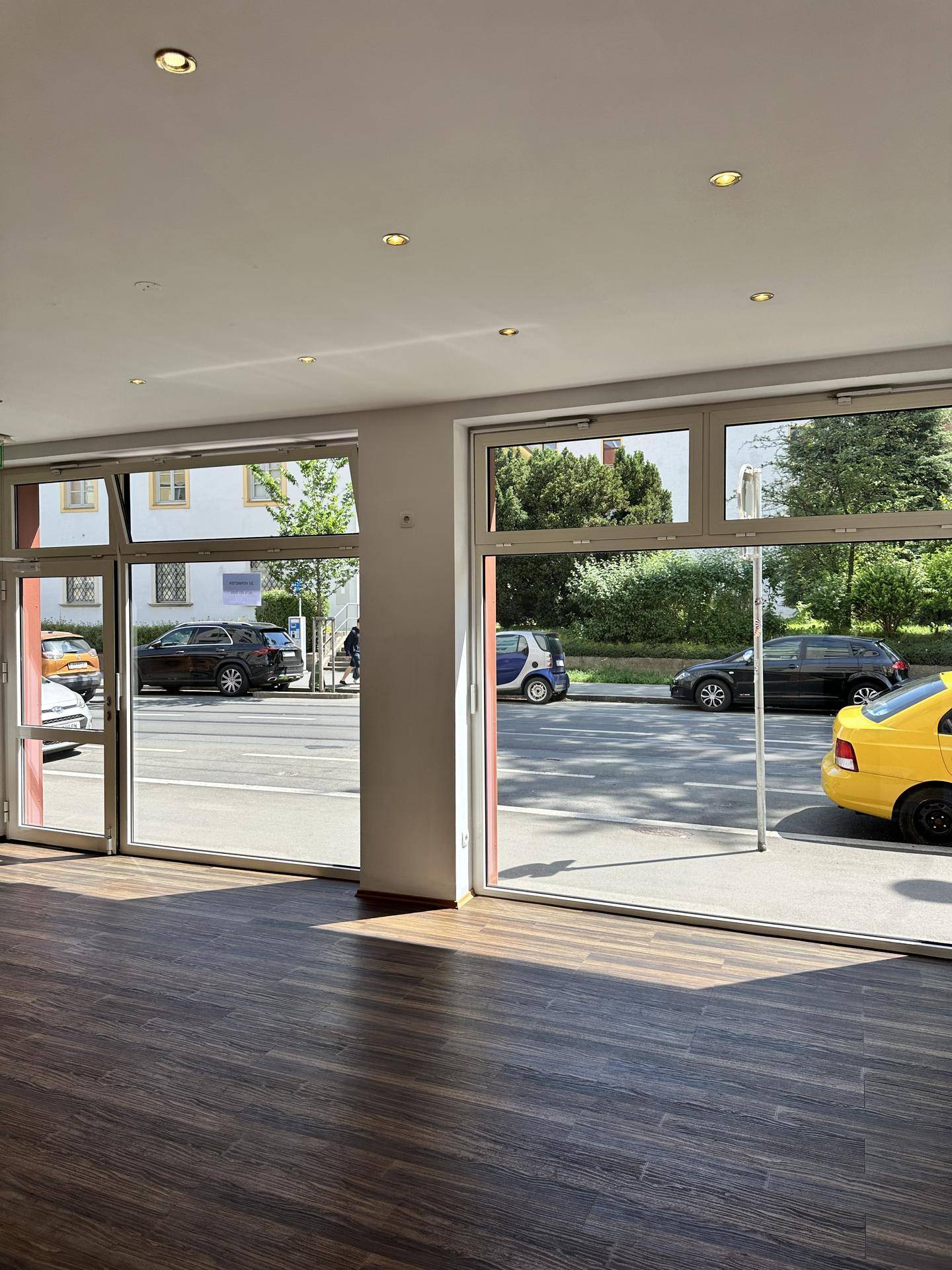 Heller Geschäftsraum mit großen Schaufenstern zur Straße, dunklem Holzboden und viel Tageslicht.