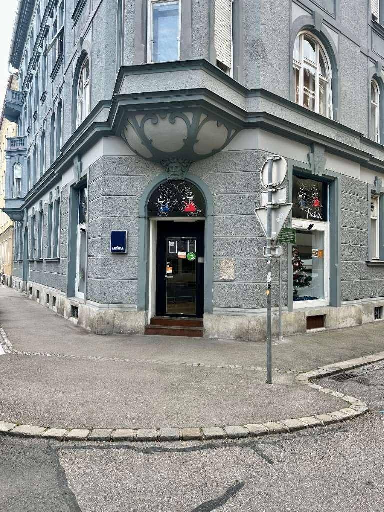 Nahaufnahme der Ecke eines grauen Gebäudes mit Schaufenstern und einem Eingangsbereich.