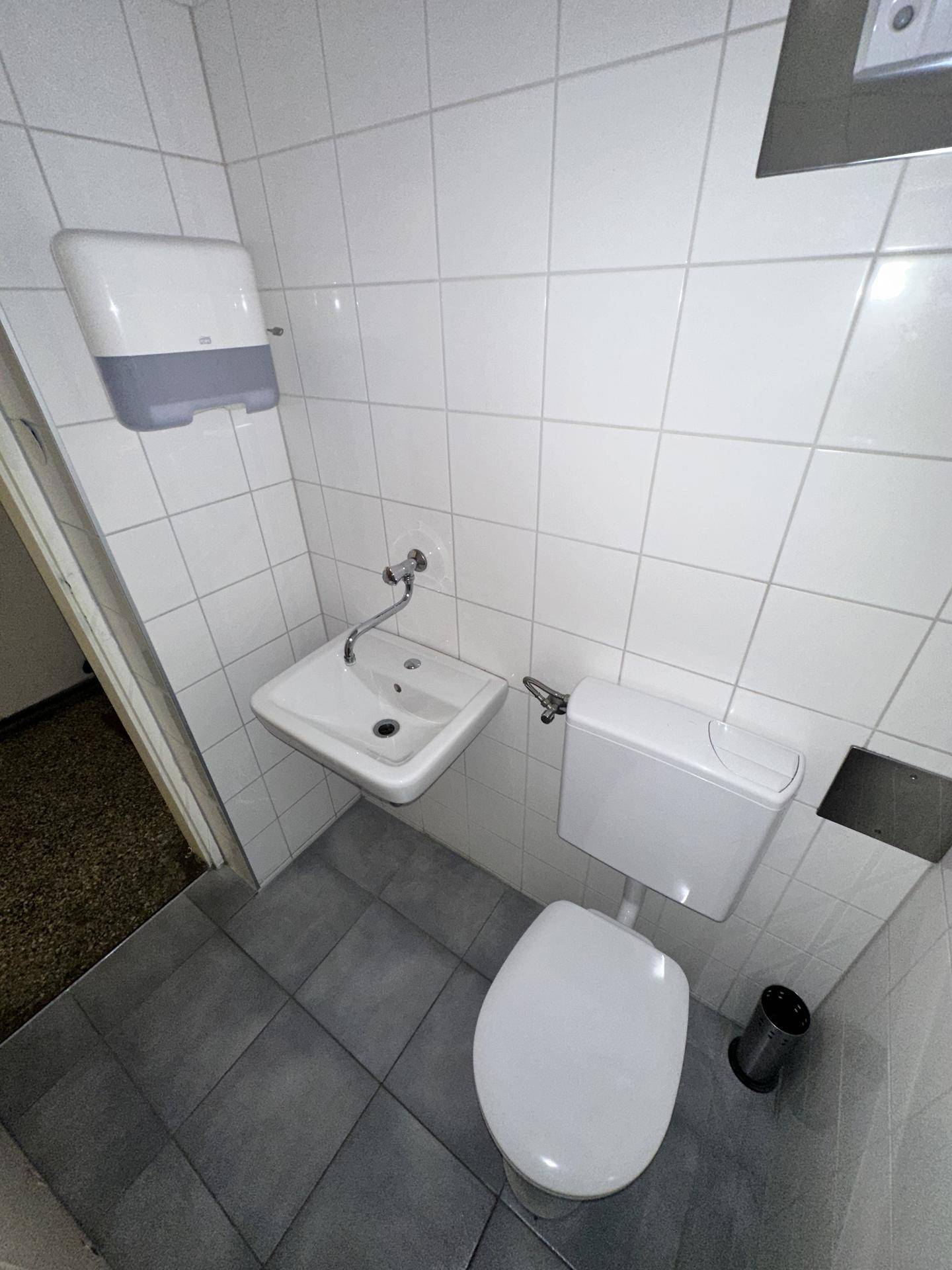 Nahaufnahme eines Toilettenraums mit Waschbecken, WC und Seifenspender an der Wand.