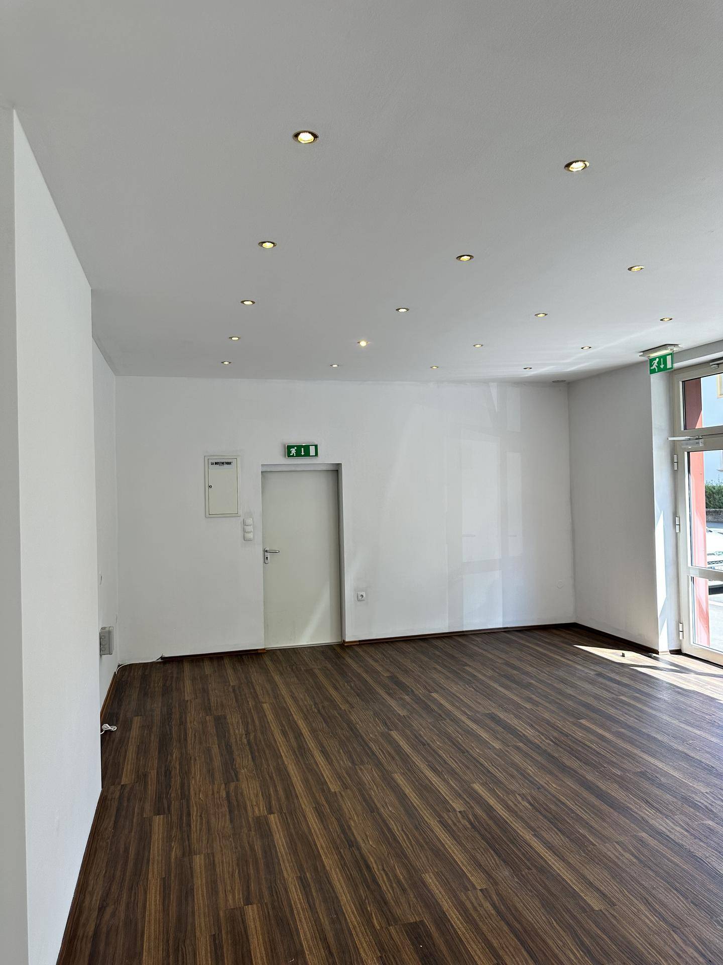 Leerer Geschäftsraum mit weißen Wänden, dunklem Holzboden und einer Tür mit Notausgangsschild.