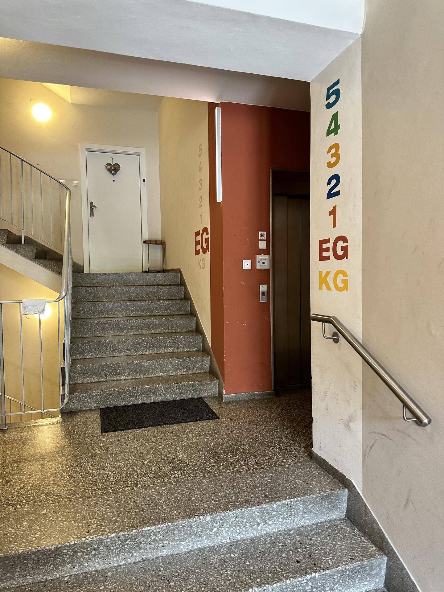 Treppenhaus mit Terrazzoboden, Handlauf und Stockwerksanzeige, inklusive Aufzugzugang.