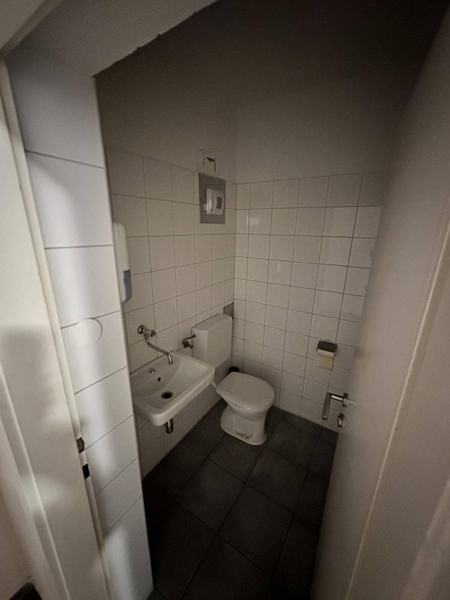 Kleiner Toilettenraum mit weiß gefliesten Wänden, Waschbecken und WC, in dunkler Beleuchtung.