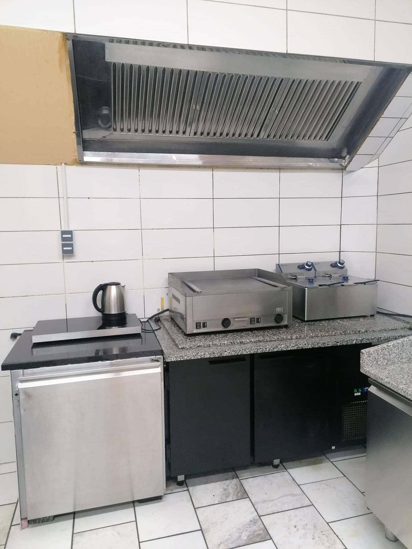 Kochbereich einer Gewerbeküche mit Dunstabzugshaube, Kochfeld und Fritteusen.
