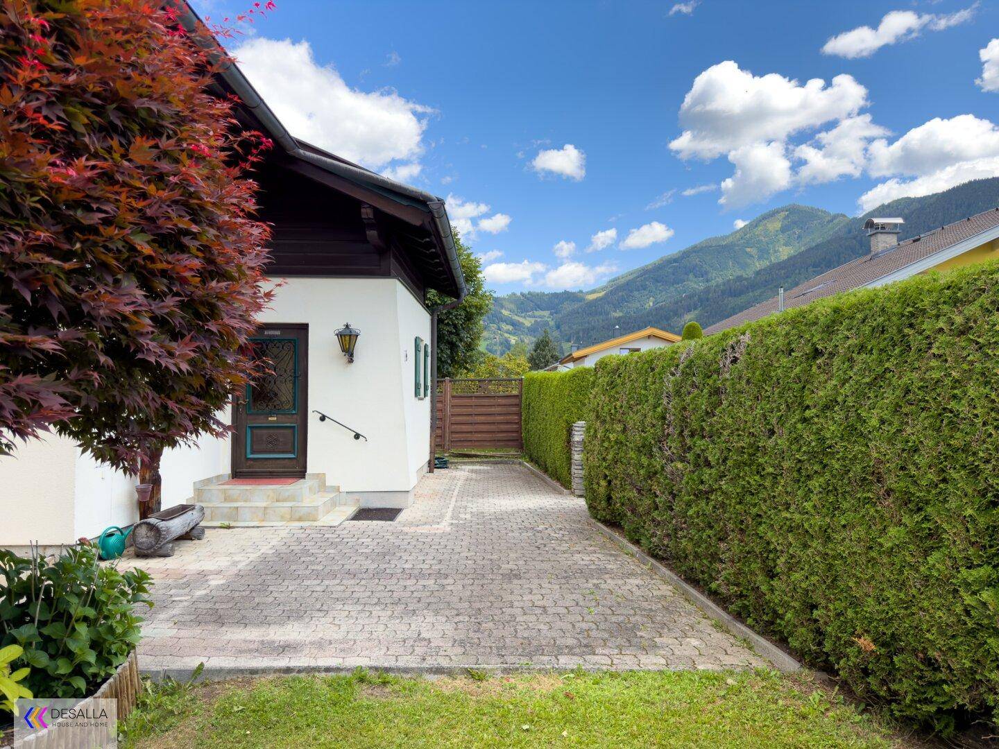 Seitenansicht des Hauses mit Eingangsbereich, gepflastertem Weg und einer hohen Hecke.