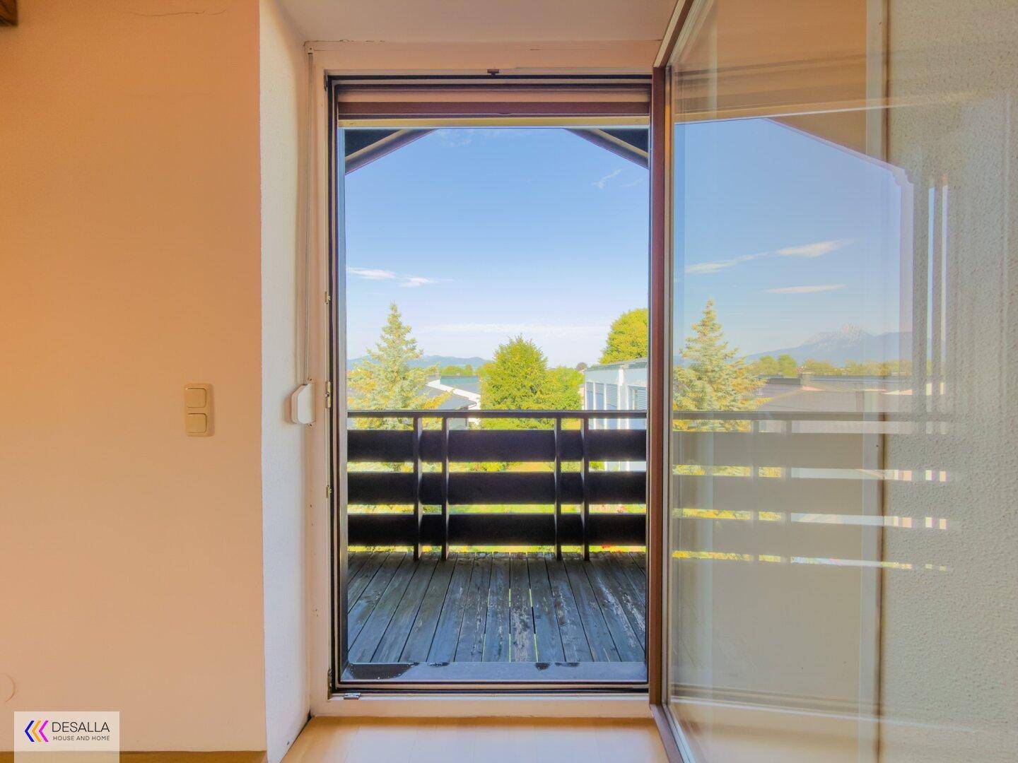 Blick vom Innenraum auf einen kleinen Balkon mit Holzboden und grüner Umgebung.