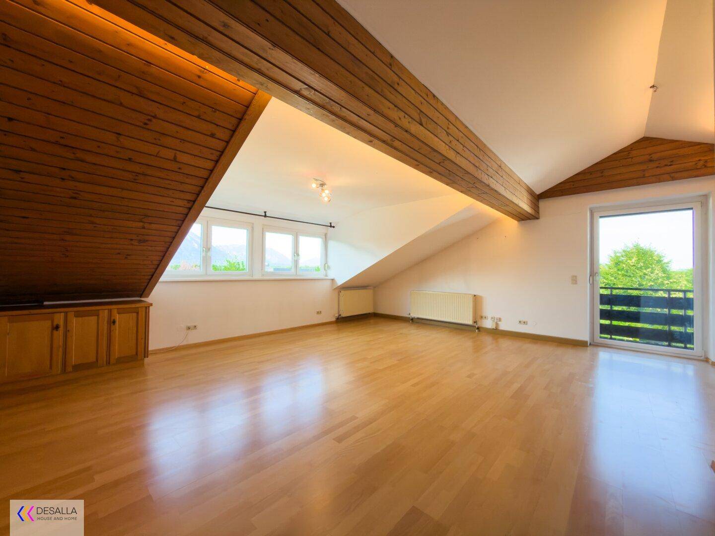 Helles Dachgeschosszimmer mit Holzbalken, Parkettboden und Zugang zu einem kleinen Balkon.