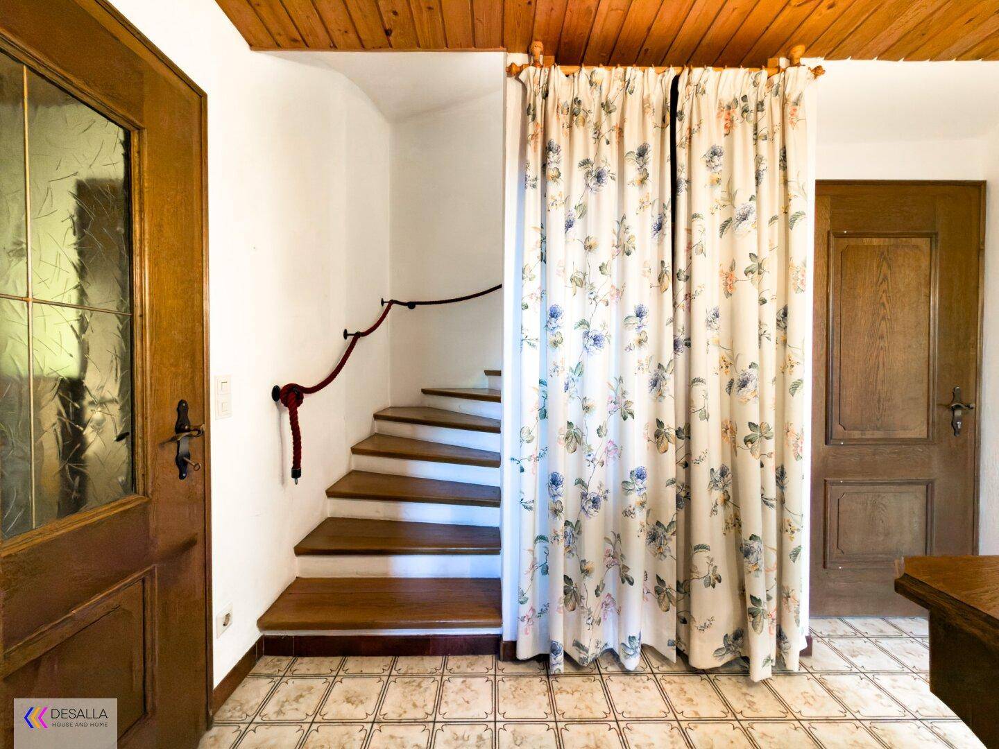 Flur mit Holztreppe, floralem Vorhang und Fliesenboden.