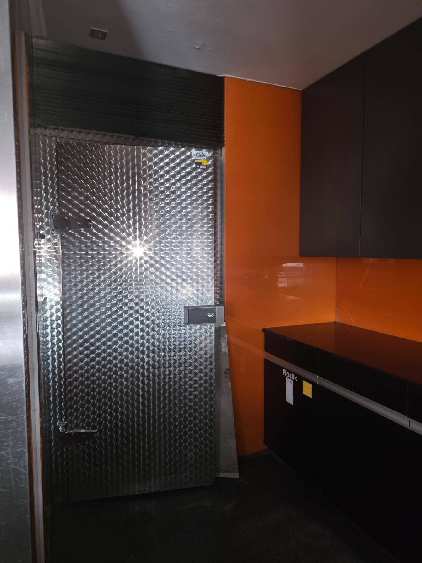 Große, strukturierte Metalltür eines Kühlraums neben orangefarbenen Wandpaneelen in einem funktionalen Bereich.