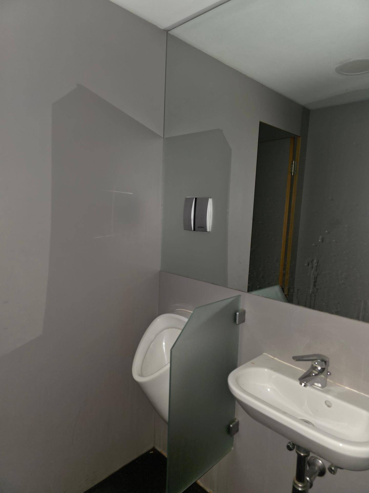 Badezimmerbereich mit Urinal, kleiner Trennwand und Waschbecken, in einem schlichten, funktionalen Design.