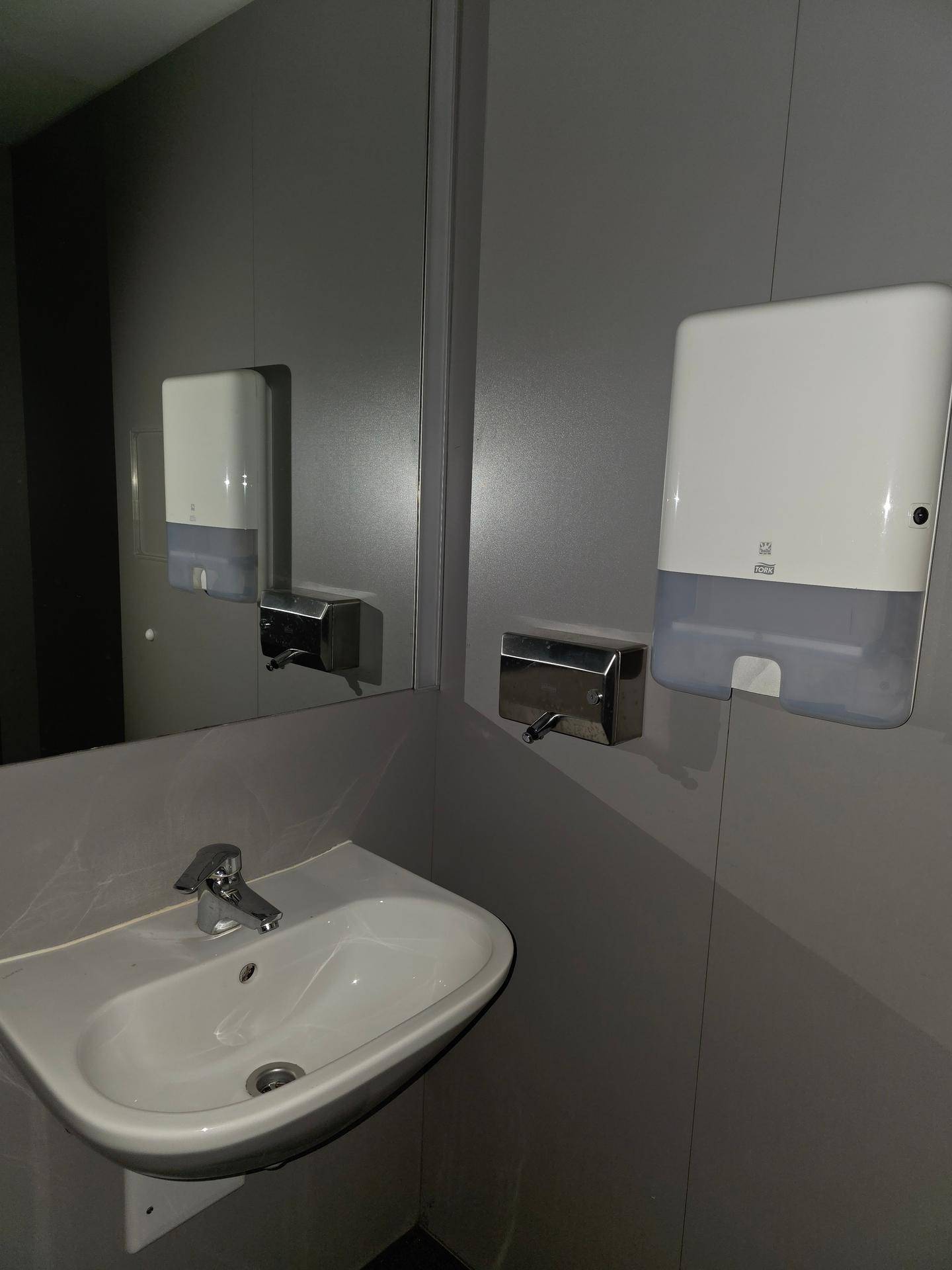 Funktionales Badezimmer mit Waschbecken, Spiegel und Spendern für Seife und Papierhandtücher.