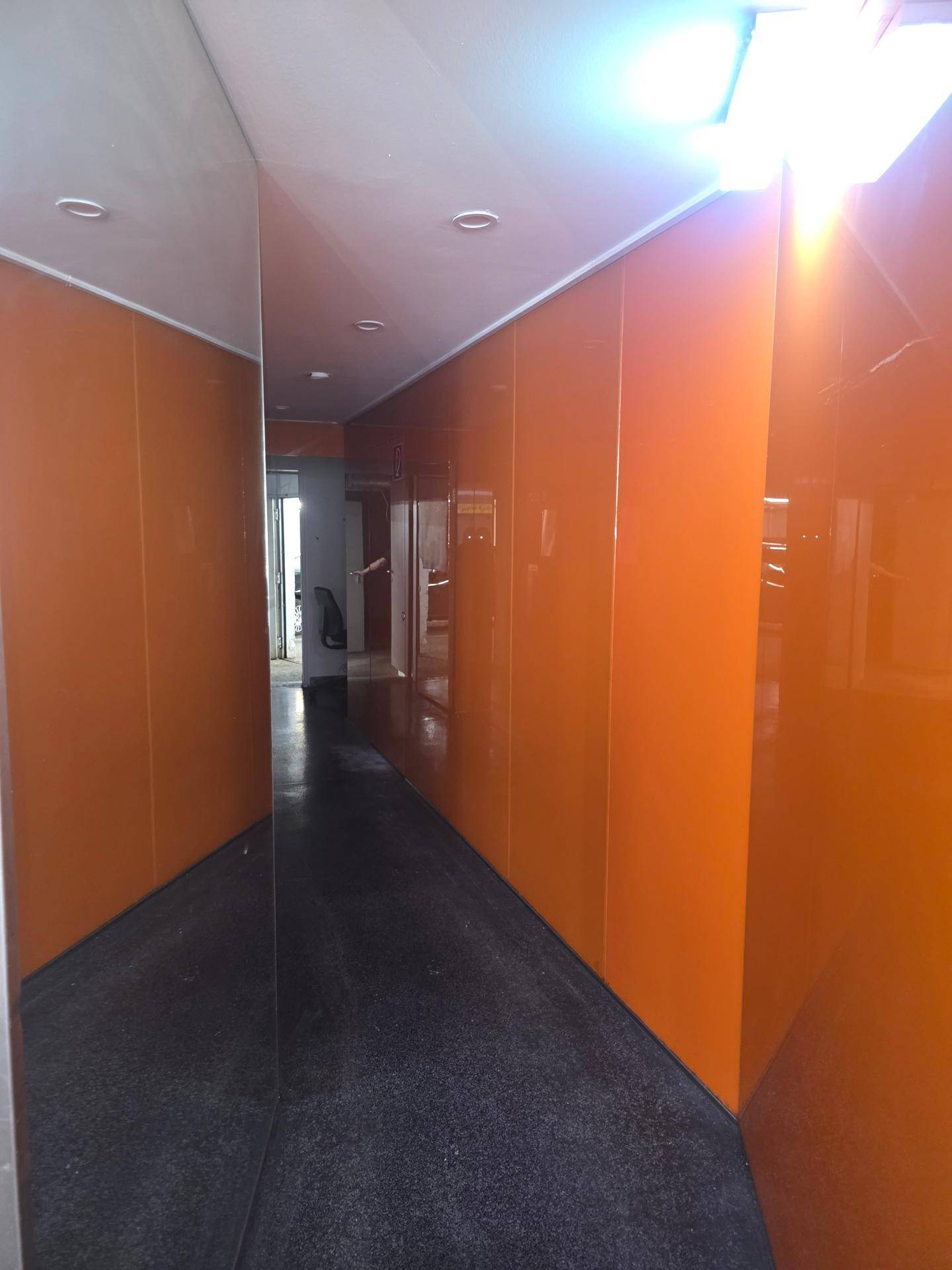 Langer Flur mit leuchtend orangefarbenen Wandpaneelen und einer reflektierenden Oberfläche, die den Raum optisch erweitert.