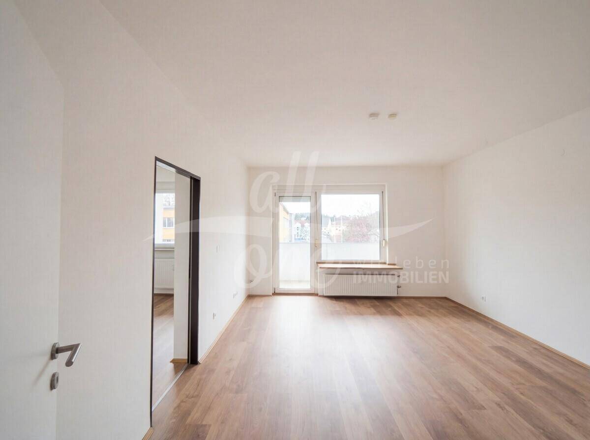 Helles, leeres Wohnzimmer mit Laminatboden und Zugang zum Balkon.