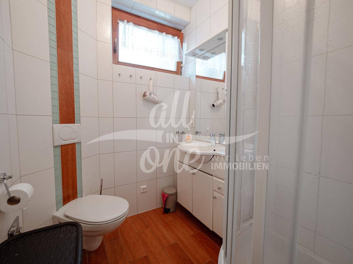 Helles Badezimmer mit weißen Fliesen, WC, Waschbecken, Dusche und einem Fenster für Tageslicht.