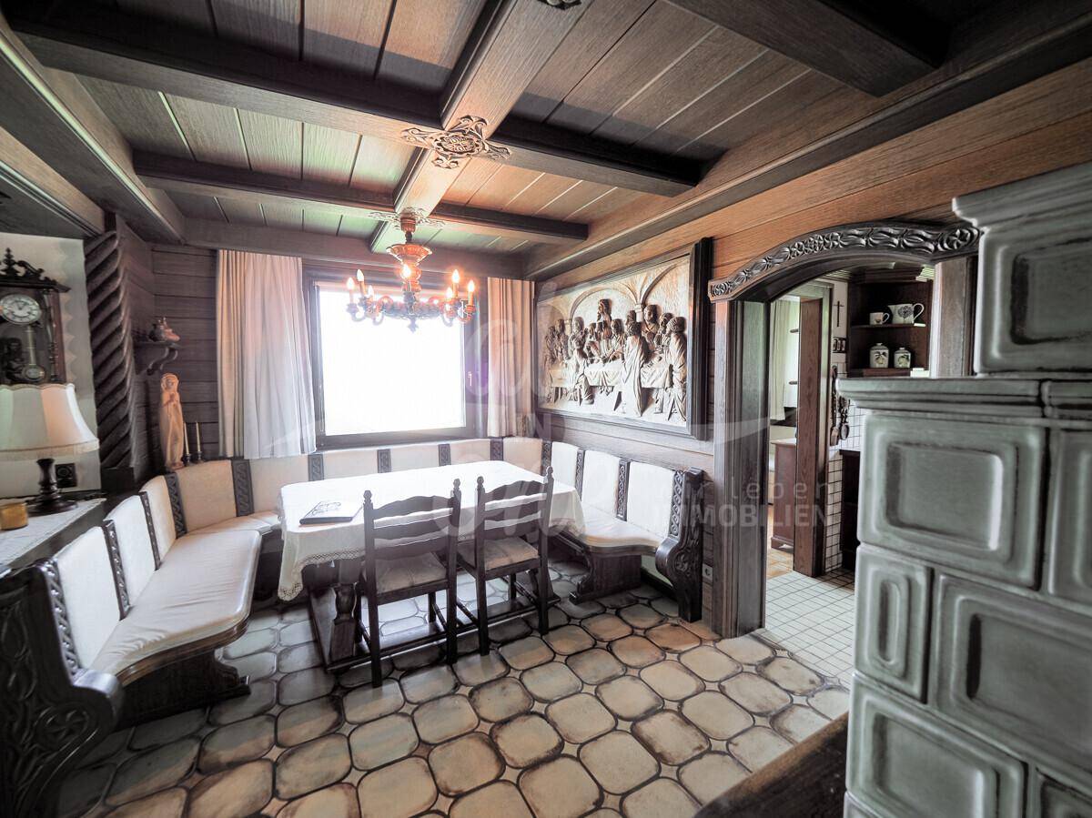 Rustikaler Essbereich mit Kachelofen, Holzbalkendecke und traditionellen Eckbänken am Fenster.