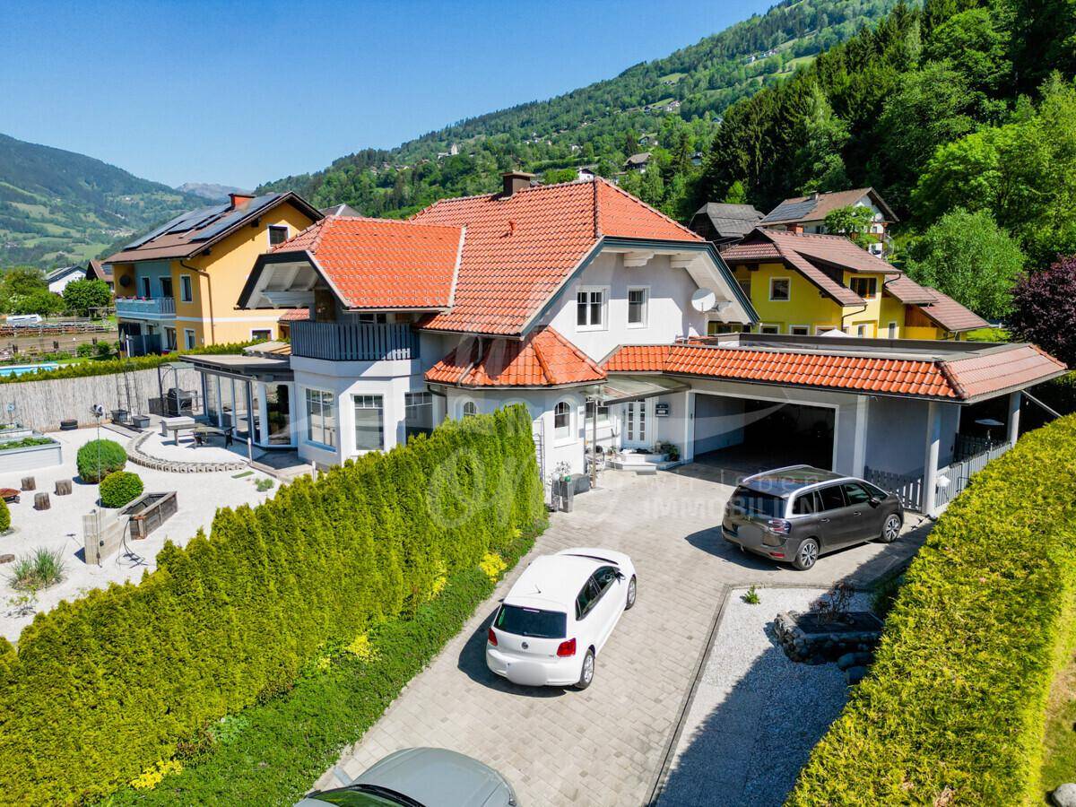 Luftaufnahme eines Einfamilienhauses mit gepflegter Auffahrt, umgeben von grünen Hecken und Bergen.