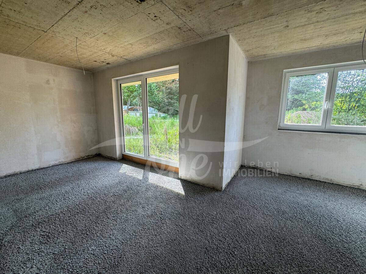 Rohbau-Zimmer mit großer Schiebetür zum Balkon und Blick auf die grüne Umgebung.