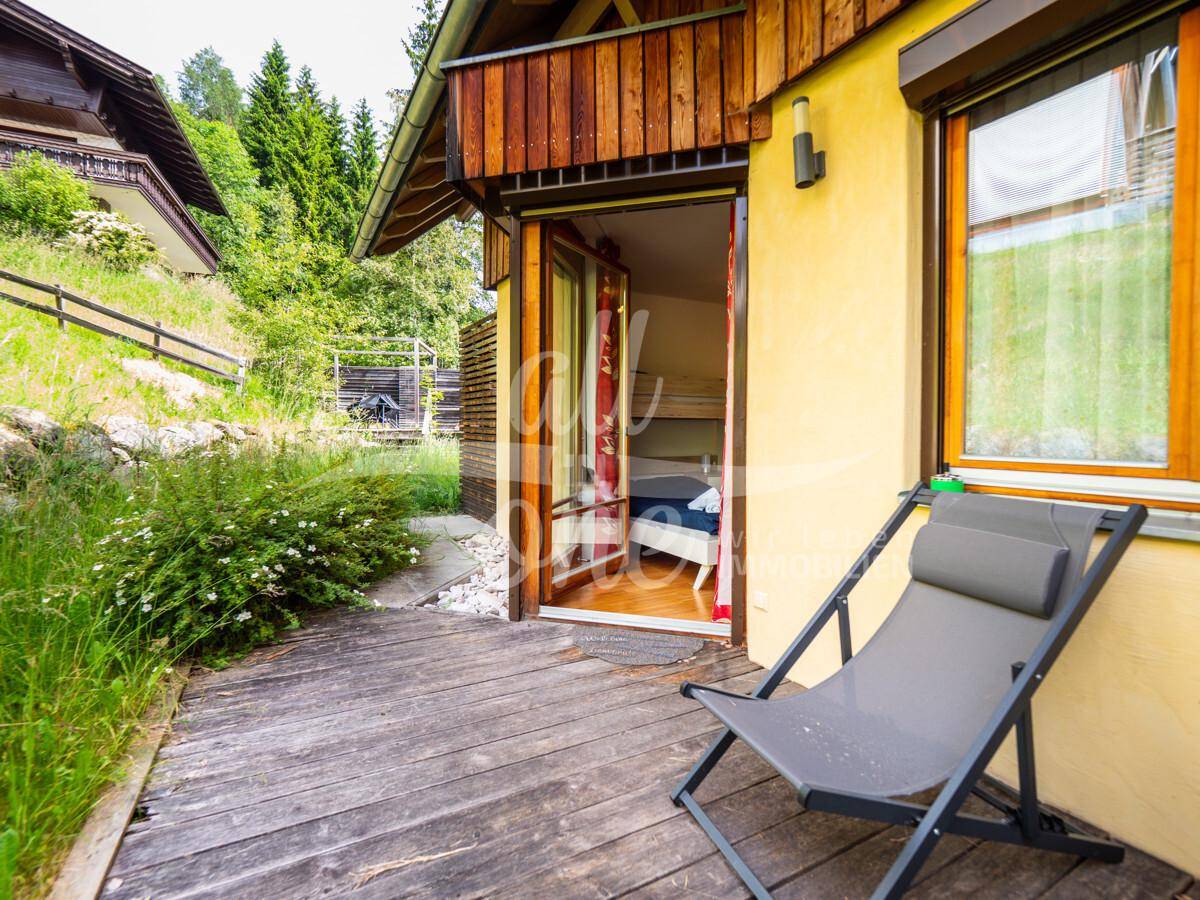 Holzterrasse mit Liegestuhl und direktem Zugang zum Innenbereich, umgeben von grüner Landschaft.