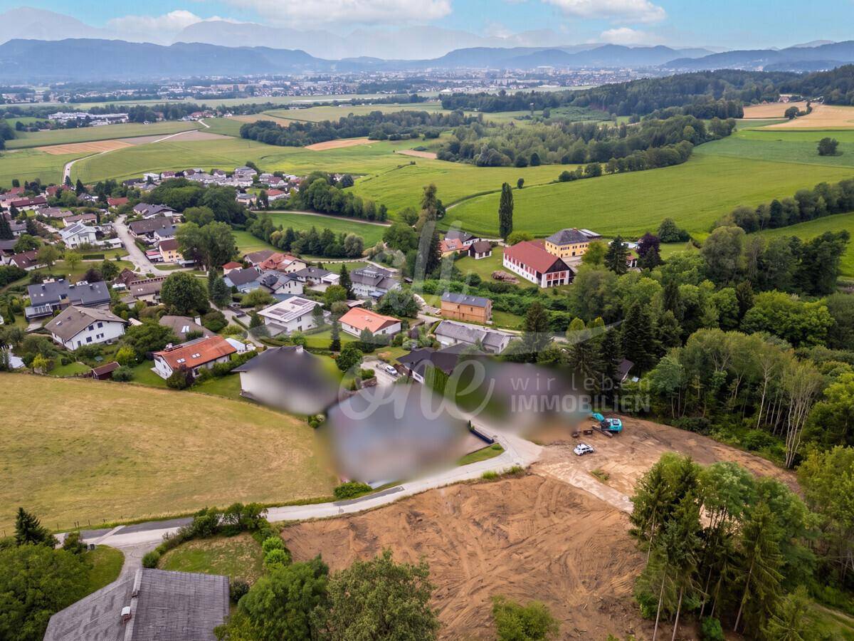 Panoramablick aus der Luft auf ein Baugrundstück, umgeben von grünen Feldern und einer Wohnsiedlung.