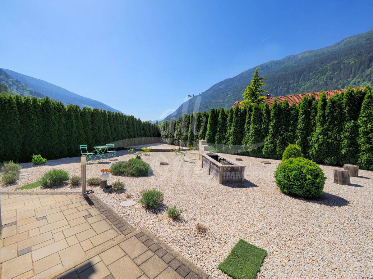 Gepflegter Gartenbereich mit Kiesflächen, Hochbeeten und hohen Hecken vor Bergkulisse.