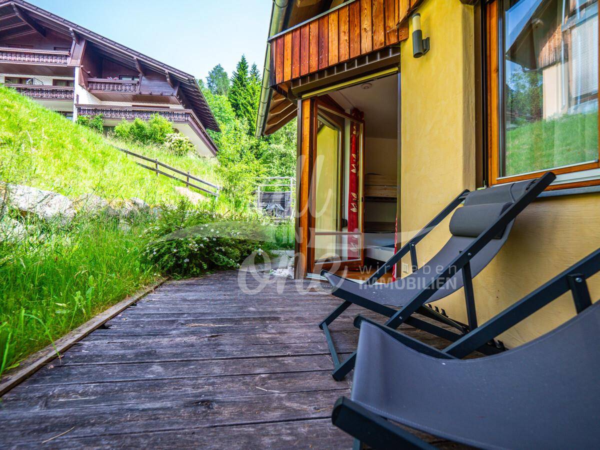 Holzterrasse mit Liegestühlen und direktem Zugang zu einem Innenraum, umgeben von Natur.