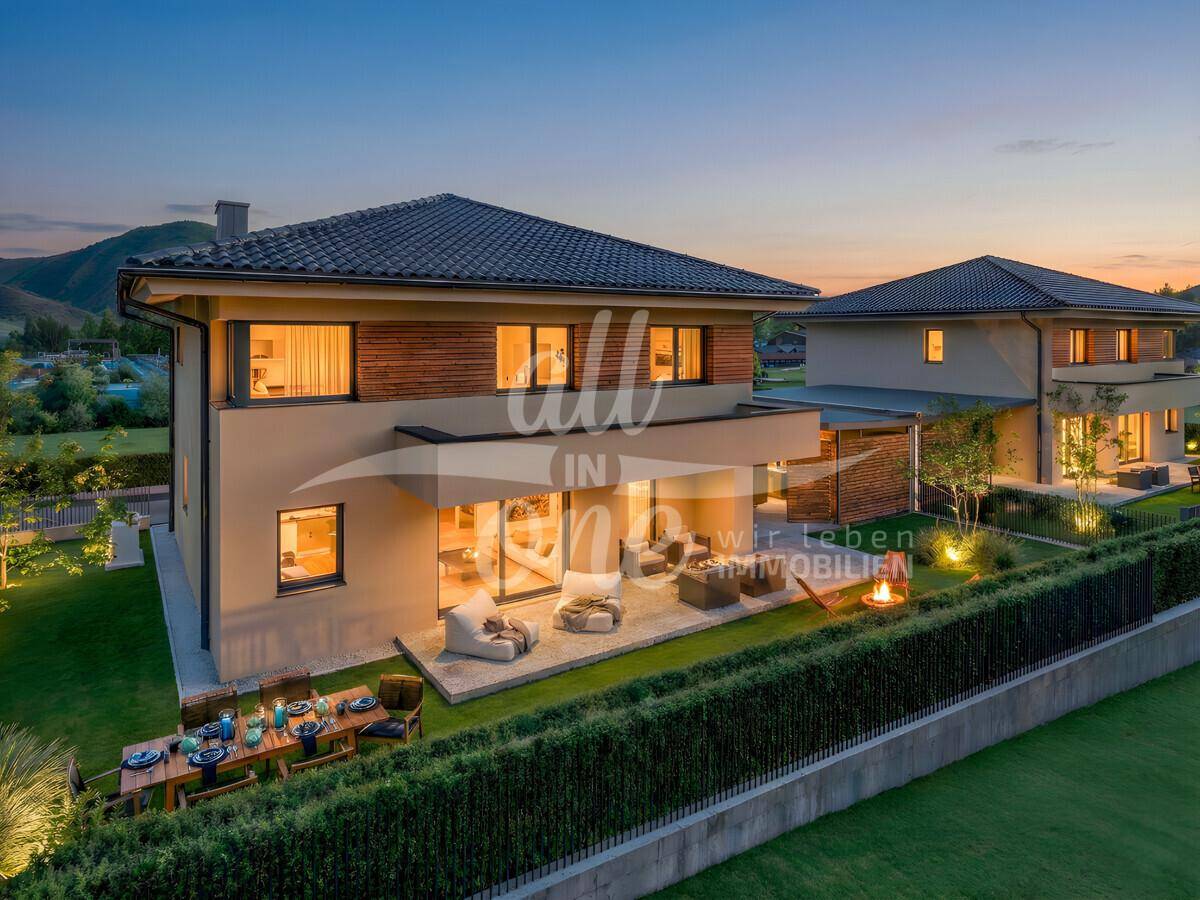 Abendliche Ansicht des modernen Hauses mit beleuchtetem Garten, Terrasse und gemütlicher Feuerstelle.