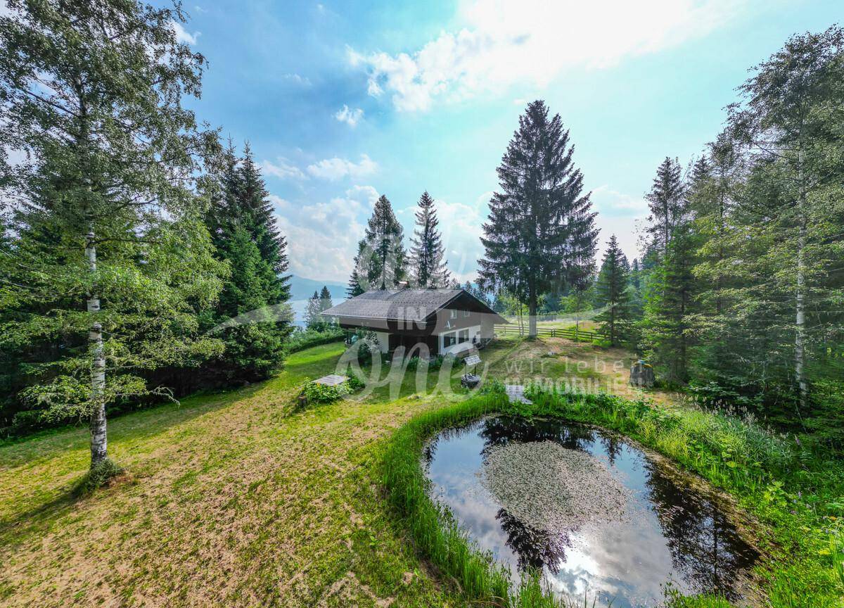 Idyllisches Haus mit einem kleinen Teich und weitem Blick auf die umliegende Berglandschaft unter blauem Himmel.