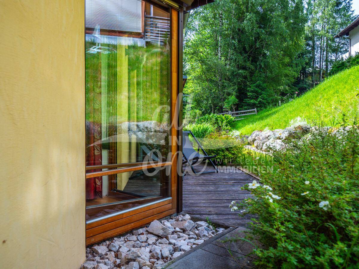 Zugang zu einem Innenraum über eine Glastür, mit einer Holzterrasse und üppigem Grün.